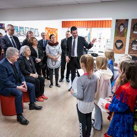 La gouverneure générale Mary Simon et un homme sont assis sur un banc rouge. Des étudiants se tiennent devant eux, discutent et tiennent une feuille de papier. Quelques personnes sont également debout derrière eux.