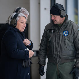 La gouverneure générale Simon est debout à l’extérieur et écoute un homme parler. Ils portent tous les deux des écouteurs. Deux personnes sont debout derrière eux.