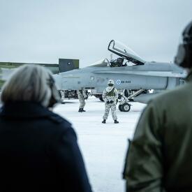 La gouverneure générale Mary Simon est debout à l'extérieur à côté d'un homme. Ils portent des écouteurs. Une personne est debout à l'arrière-plan devant un avion.