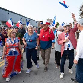Governor General Mary Simon and Mr. Whit Fraser are walking with a crowd of people outside, participating in National Acadian Day festivities.