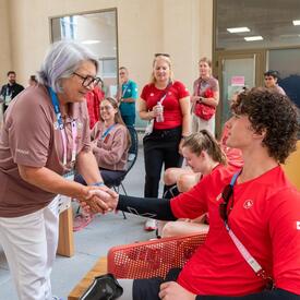 The Governor General of Canada Mary Simon shake hands with a seated Team Canada athlete. Several people, including other athletes and officials, are smiling and observing the interaction.
