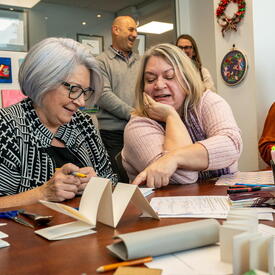 Governor General Mary Simon sitting next to a woman at a table full of craft supplies. The woman is pointing something out to her.