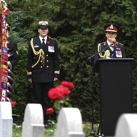 The Governor General of Canada, in a canadian army uniform, is speaking at a podium, in a cemetary during a Remembrance Day Commemoration in Belgium. Her female Aide-de-camp is standing on her right side.