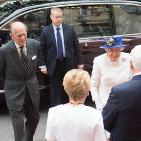 Her Majesty the Queen and the Duke of Edinburgh are greeted by Governor General Johnston and Mrs. Sharon Johnston. A bodyguard in a suit stands next to a vehicle in the background.