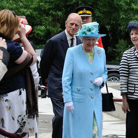 The Queen and the Duke of Edinburgh walk outdoors, along a sidewalk beside a green lawn. The Queen waves. They are accompanied by several men in suits and military uniforms.