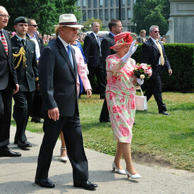 The Queen and the Duke of Edinburgh walk outdoors, along a sidewalk beside a green lawn. The Queen waves. They are accompanied by several men in suits and military uniforms. 