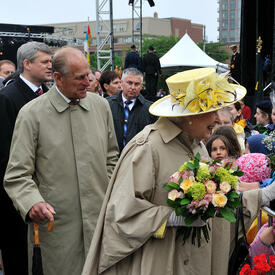 Her Majesty The Queen smiles as she receives a bouquet from a person in a crowd standing behind a barrier. The Queen holds more flowers in her other hand. The Duke of Edinburgh and Prime Minister Stephen Harper follow along behind her. 