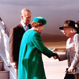 The Queen shakes hands with Governor General Sauvé, who curtsies. The Duke of Edinburgh is standing behind the Queen. A uniformed officer salutes in the background. All are standing at the bottom of a set of stairs extending from an airplane onto the tarm