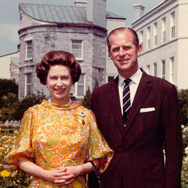 The Queen and the Duke of Edinburgh stand in the private gardens at Rideau Hall. They are both smiling. The Queen is wearing a yellow-and-orange-patterned dress; the Duke is wearing a dark suit.