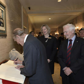 The Duke of Edinburgh writes in a large book as Governor General David Johnston looks on. The pair are in a lobby with other people.