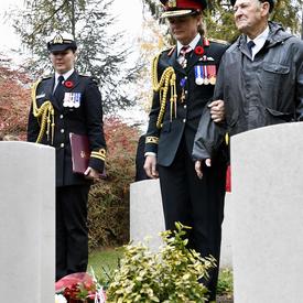 La gouverneure générale du Canada, en uniforme de l'armée canadienne, s'arrête devant une tombe dans un cimetière avec un ancien combattant à ses côtés.