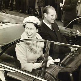 The Queen and the Duke of Edinburgh smile at the crowd while riding in an open-air vehicle. The photo is black and white.