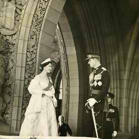 La reine, vêtue d’une robe de bal blanche classique et d’une écharpe en fourrure, se tient face au duc d’Édimbourg, qui porte un uniforme militaire. Le couple se tient sous une arche de pierre sculptée de motifs raffinés.