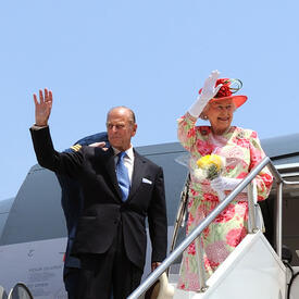 The Queen and the Duke of Edinburgh wave from the top of a white staircase. They are standing in front of the open door of an airplane.