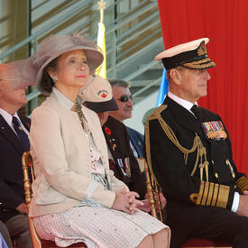 La Reine, la gouverneure générale Clarkson et le duc d’Édimbourg sont assis côte à côte lors d’un événement à l’extérieur. D’autres dignitaires et invités sont assis derrière eux.