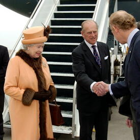 The Duke of Edinburgh shakes hands with John Ralston Saul as The Queen and Governor General Clarkson look on. The group is standing on the tarmac at an airport, at the bottom of a set of stairs leading up to an airplane.