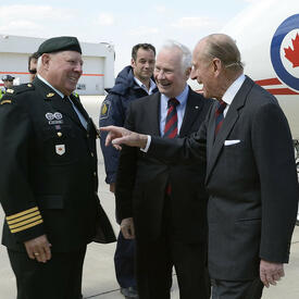 The Duke of Edinburgh laughs as he speaks with Colonel Joe Aitchison, with Governor General David Johnston looking on. The men are standing on the tarmac outside of an airport. A plane bearing the logo of the Canadian Air Force is in the background.