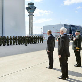 The Duke of Edinburgh and Governor General Johnston stand on the tarmac at an airport. They are facing a group of military members dressed in uniform and standing in formation. On their right, Colonel Joe Aitchison directs the group with an epée in hand. 