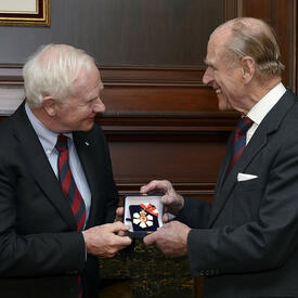 Le gouverneur général David Johnston remet au duc d’Édimbourg une petite boîte contenant l’insigne de l’Ordre du Canada. L’insigne a la forme d’un flocon de neige blanc stylisé avec une feuille d’érable rouge en son centre.