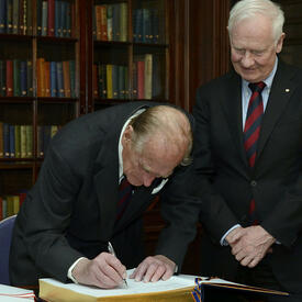 The Duke of Edinburgh signs a large book with gold-leaf edges as Governor General Johnston looks on.