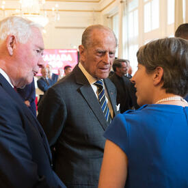 His Royal Highness The Duke of Edinburgh and Governor General David Johnston speak to a woman. They are in a large reception area with other guests in the background. A chandelier hangs from the ceiling.