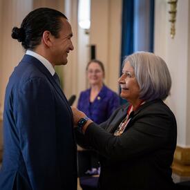 Governor General Simon is smiling to Premier Wab Kinew while pining a Coronation Medal on his vest.