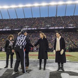 Governor General of Canada Julie Payette shakes hands with a referee at the 2018 Grey Cup Championship, in the centre of the field of the Commonwealth Stadium in Edmonton. The stadium is filled with fans.