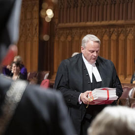 A senator approaches His Excellency with documents in hand.
