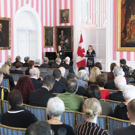 The Governor General, Julie Payette, stands at a podium and delivers a speech to a seated crowd of around 200 people. 