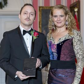 Michaël Trahan stands next to Governor General Julie Payette.  They hold a leather bound book and smile at camera. 