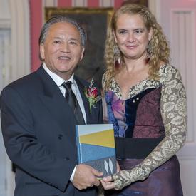 Darrel J. McLeod  stands next to Governor General Julie Payette.  They hold a leather bound book and smile at camera