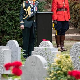 Governor General Julie Payette stands at a podium.  In the foreground are white tomb stones.  Behind her, to her right, a mountie in red surge stands at attention. 