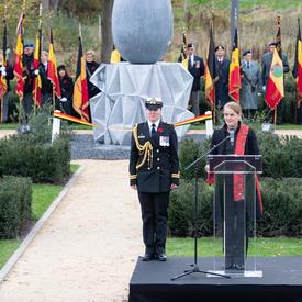 The Governor General of Canada is speaking at a podium, with her female Aide-de-camp on her right side. A giant concret sculpture in the shape of a teardrop is behind her.