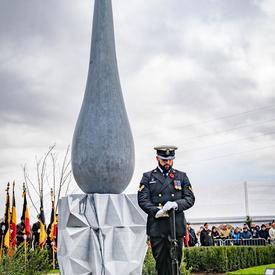 A serviceman stands in front of a monument, eyes towards the ground.  The monument's base has an angular triangle pattern, the top is the shape of a large teardrop. 