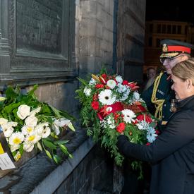 The Governor General of Canada is laying a wreath of flower at a wall monument at night with the help of a military member.