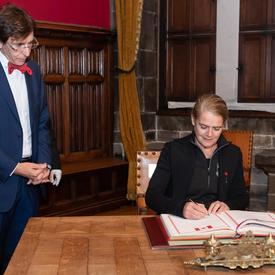 The Governor General is signing a guestbook. The Mayor of Mons, Elio di Rupo, is standing at her right is watching her.