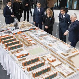 The Governor General is looking at a miniature model on a large rectangular table. A gentleman on her left is explaining the model as he points to it.  They are accompanied by half a dozen other people.  