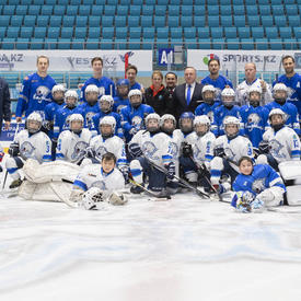 Two youths’ hockey teams pose on the ice in an arena. The children are wearing white and blue jersey and full hockey gear. In the background stand adults, including the Governor General, Julie Payette. 