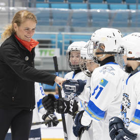 The Governor General, Julie Payette, shakes the hands of children dressed in hockey gear.  They are on the ice in an arena. 