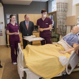 Her Excellency stands around a hospital bed where students are caring for a mannequin. 