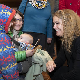 The Governor General, Julie Payette, is squatting down and speaks to a child wearing a winter coat. 