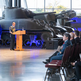  The governor general stands in front of a podium and addresses the crowd, largely in military uniform.  A tank is   behind her, in the background. 