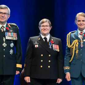  Recipient Chief Petty Officer 2nd Class Marie Nathalie Isabelle Scalabrini stands between the Vice Chief of the   Defence Staff, Lieutenant-General Paul Wynnyk (on the left) and Her Excellency the Right Honourable Julie Payette   (on the right).