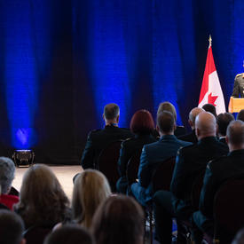 The Vice Chief of the Defence Staff, Lieutenant-General Paul Wynnyk stands in front of a podium and addresses the   crowd, largely in military uniform.  Her Excellency the Right Honourable Julie Payette is seated and is listening   to the speech.