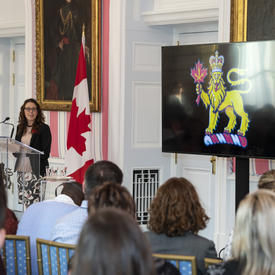 A woman speaks at a podium in front of a group of people sitting on chairs in front of her. A large TV screen beside her shows the vice-regal lion symbol.