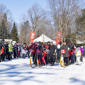 There is a large crowd standing outdoors in winter.  In the center of the photo are two rows of people strapped to large multi-persons cross-country skis.  They are at a start line getting ready to being a friendly race. 