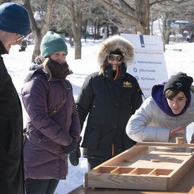 A young man wearing a tuque and a winter coat plays a game with wooden pieces.  Her Excellency stands behind him, smiling, in her winter coat. 