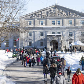 The front façade of Rideau Hall. In front of the residence is a large group of people all dressed in winter gear, walking along the main path.