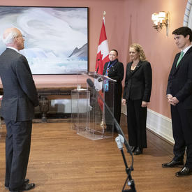 Michael Wernick, greffier du Conseil privé, se tient devant la gouverneure générale Julie Payette et le premier ministre Justin Trudeau.   Dans le coin de la salle, le maître de cérémonie se tient derrière un podium.