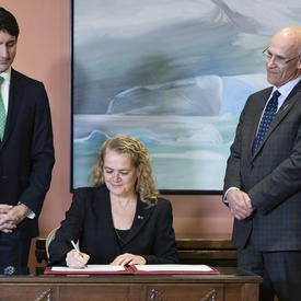 Governor General Julie Payette sits at a desk and signs a register.  Behind her, Prime Minister Justin Trudeau, on her right, and Michael Wernick, Clerk of the Privy Council, on her left.